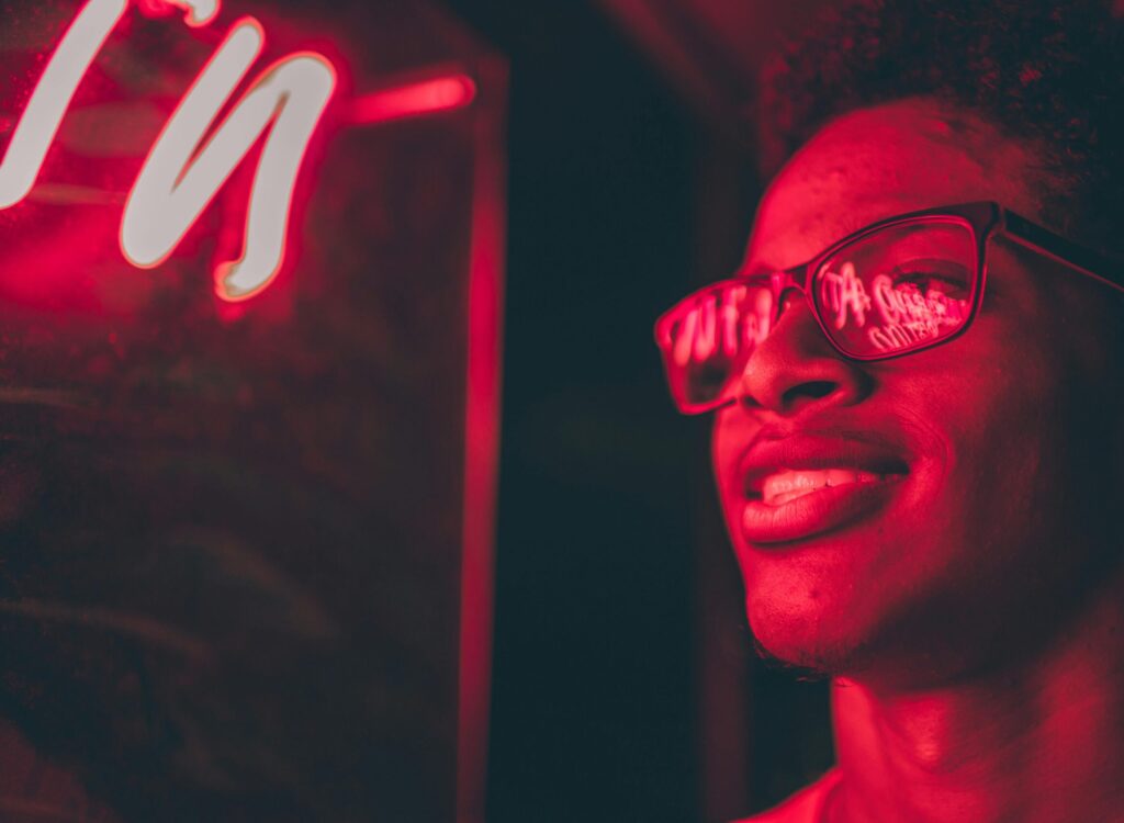 Smiling young man illuminated by vibrant neon lights, wearing reflective glasses.